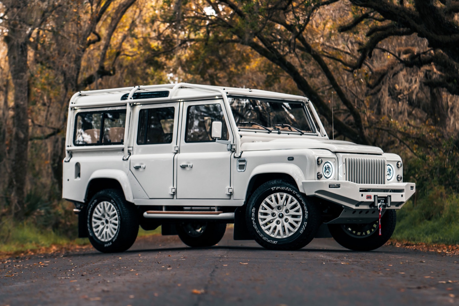A white Defender 110 restomod photographed on a live oak avenue at golden hour