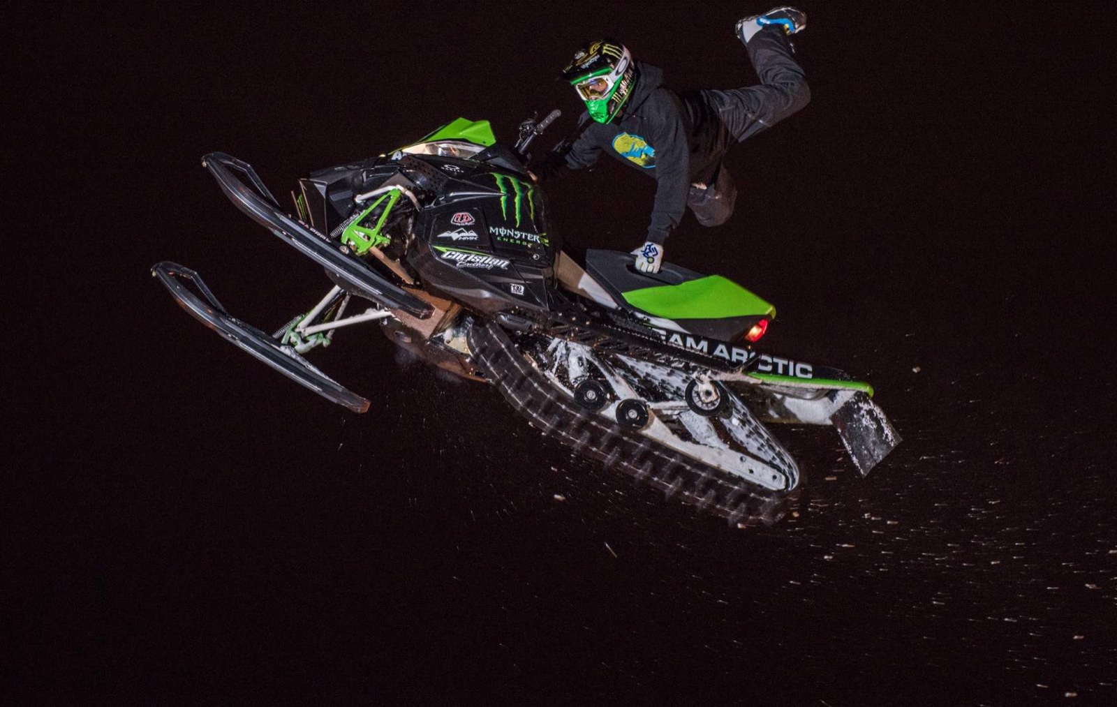 A freestyle snowmachine rider inverted mid-air against a black night sky