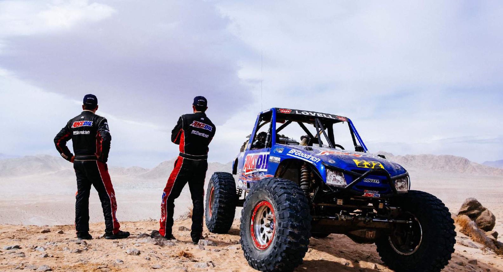 Two drivers in team suits on a desert ridge beside their off-road race car